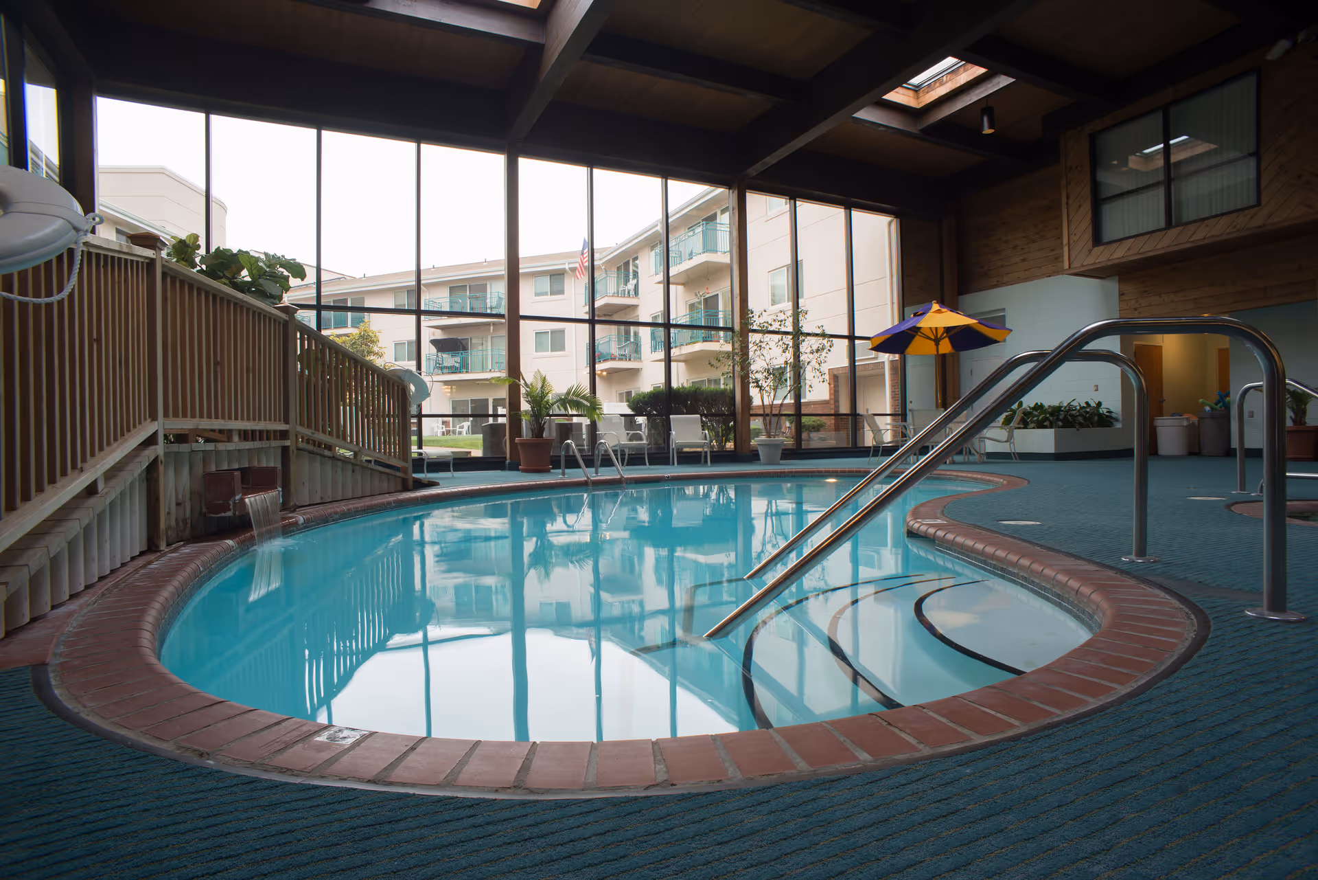 Indoor swimming pool with clear blue water, surrounded by a brick edge and blue carpeted floor. The pool area has large floor-to-ceiling windows showing an exterior view of a multi-story building with balconies. There are potted plants, a yellow and purple umbrella, and metal handrails leading into the pool.