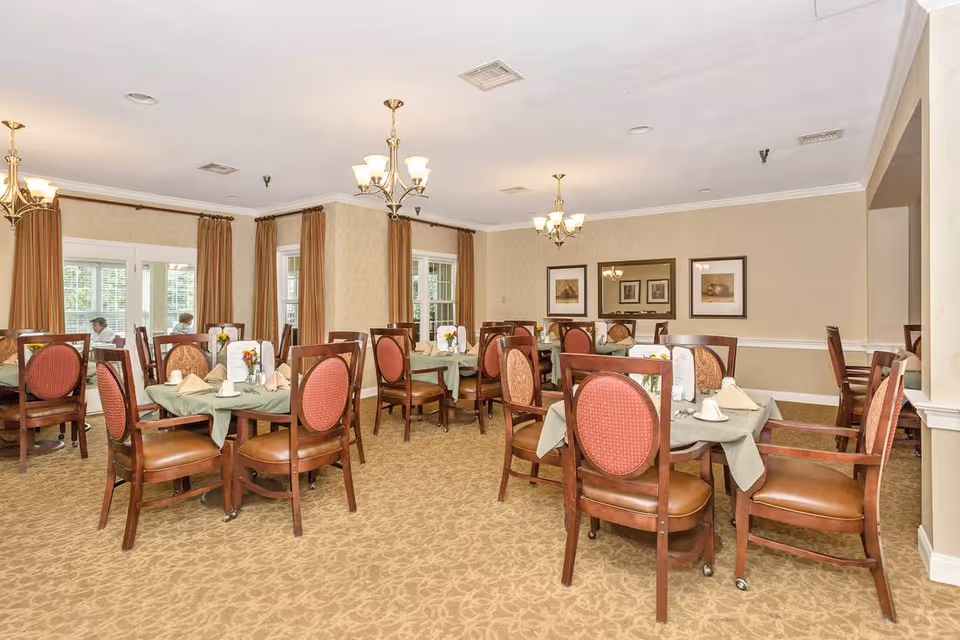 A dining room with multiple round tables covered with green tablecloths, each set with napkins, cups, and small flower arrangements. The room has beige walls, carpeted floors, large windows with brown curtains, and chandeliers hanging from the ceiling. There are framed pictures on the walls and wooden chairs with red cushioned backs and seats around the tables.