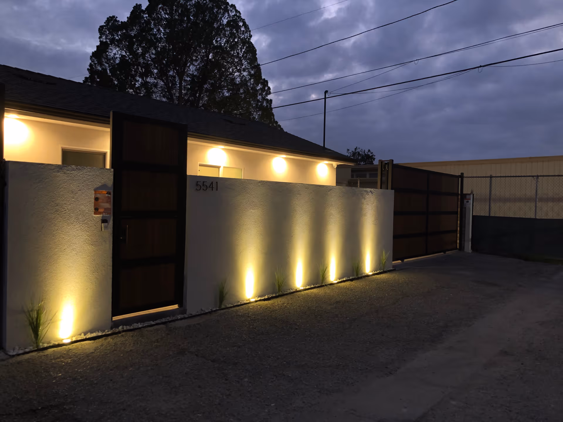 Exterior view of a building at dusk with a white wall featuring the number 5541, illuminated by ground lights. There is a wooden door set into the wall and a large gate to the right. The sky is cloudy and there are trees visible behind the building.