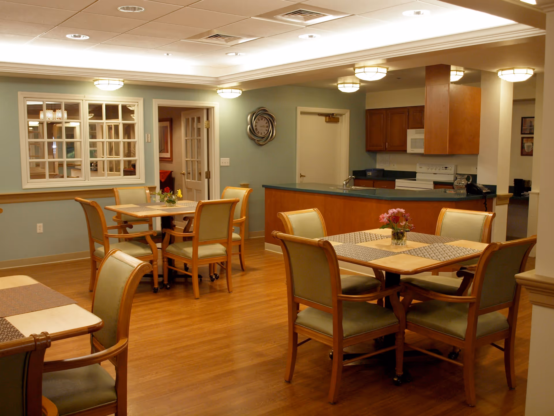 A well-lit dining area in an assisted living facility featuring wooden tables with green cushioned chairs. Each table has placemats and a small flower arrangement. In the background, there is a kitchen area with wooden cabinets, a stove, microwave, and a countertop. The walls are painted light green and there is a decorative wall clock. The floor is wooden and the ceiling has recessed lighting.