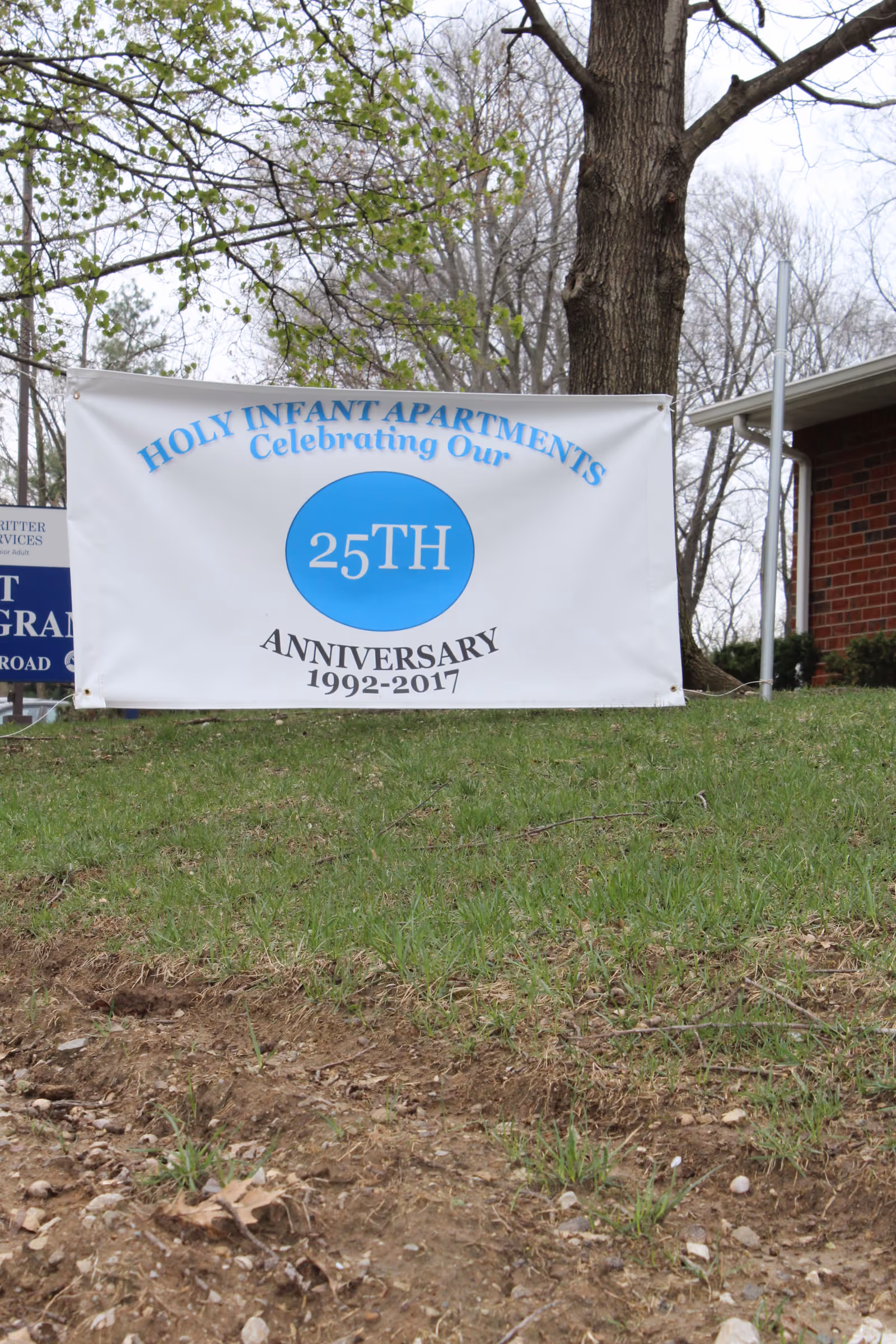 A white banner hanging outdoors on a grassy area in front of trees and a brick building. The banner reads 'Holy Infant Apartments Celebrating Our 25th Anniversary 1992-2017' with a blue circle highlighting '25TH'.