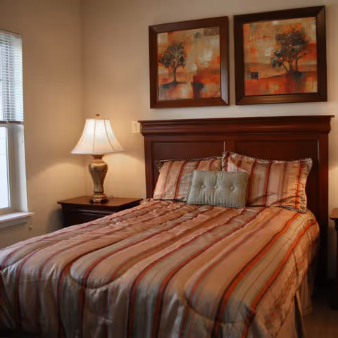 A neatly made bed with a striped comforter and pillows in a bedroom. There is a wooden headboard and two framed abstract paintings of trees hanging above it. A bedside table with a decorative lamp is next to the bed, and a window with blinds is visible on the left side.