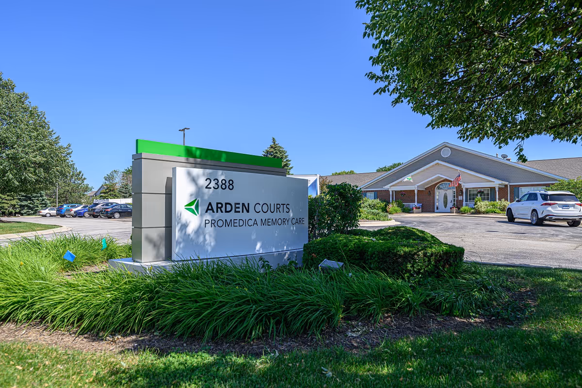 Exterior view of Arden Courts of Geneva, a memory care facility, showing the building entrance, a parking lot with cars, and a large sign with the facility name and address 2388. The scene is set on a clear sunny day with green trees and landscaping around.