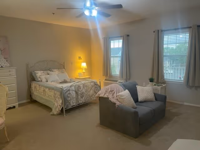 A cozy bedroom in a senior living facility featuring a metal-framed bed with floral bedding, a bedside table with a lamp, a gray loveseat with pillows and a pink throw blanket, two windows with beige curtains, and a ceiling fan with lights.
