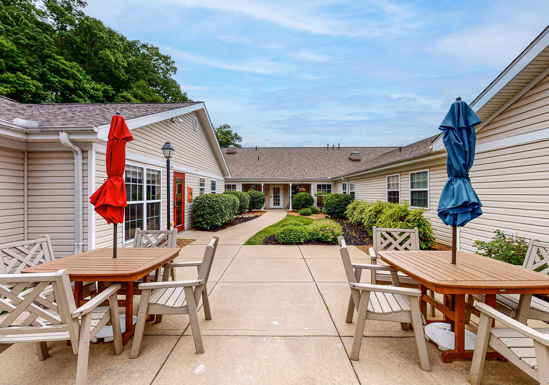 Outdoor patio area at Arden Courts - ProMedica Memory Care Community (Anderson Twp.) featuring two wooden tables with beige chairs and closed umbrellas, one red and one blue. The patio is surrounded by beige buildings with windows and green bushes, under a partly cloudy sky.