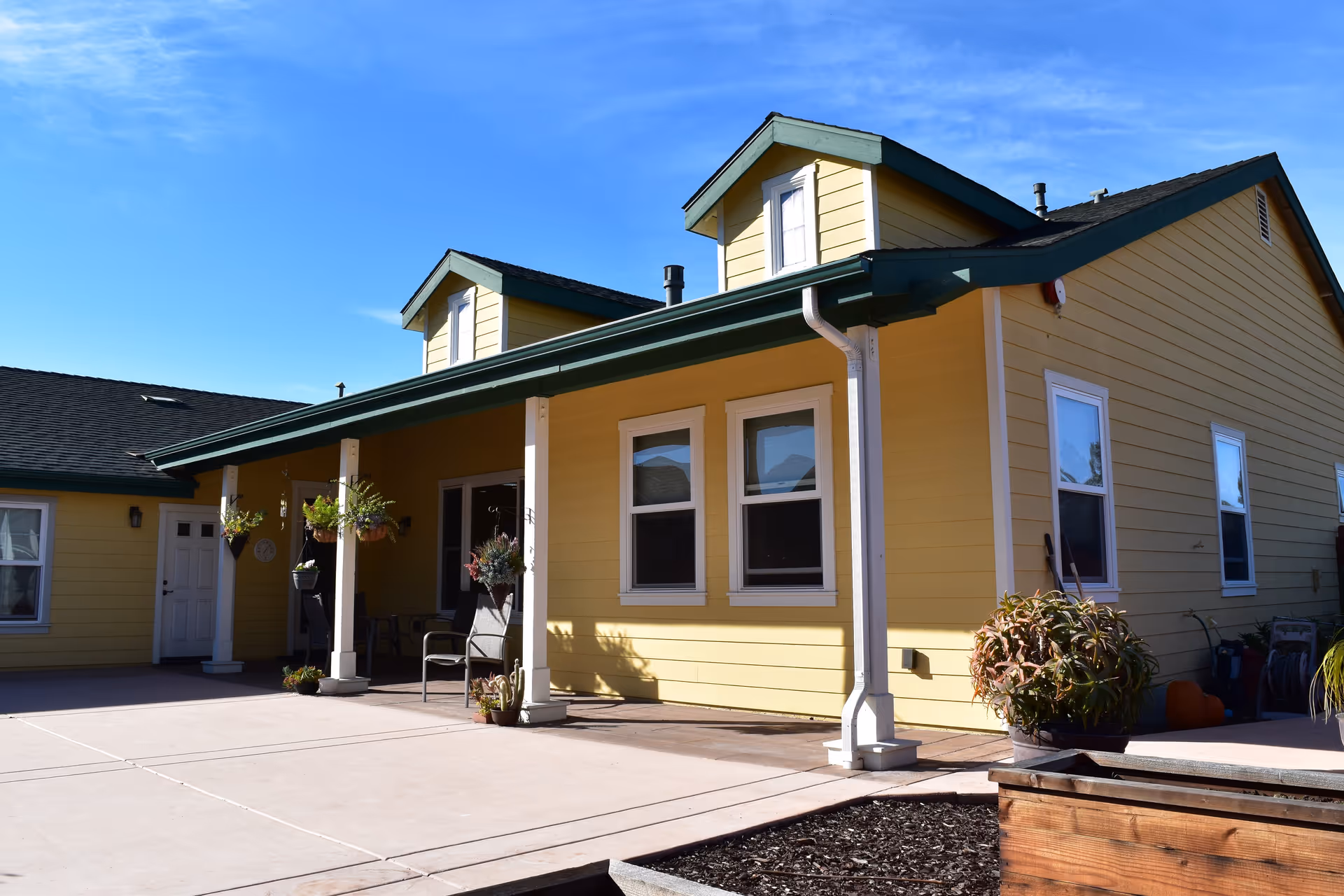 Exterior view of a yellow single-story building with green trim and a covered porch area. The porch has hanging plants and chairs, and there are several windows visible on the building. The sky is clear and blue.
