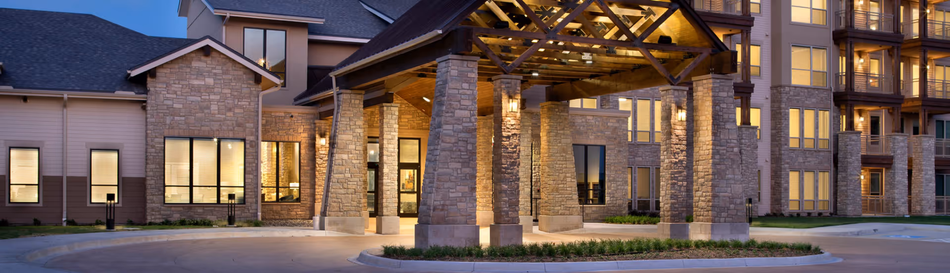 Exterior view of Stevenson Oaks senior living facility at dusk, showing a large covered entrance supported by stone pillars with warm lighting and multiple windows on the building.