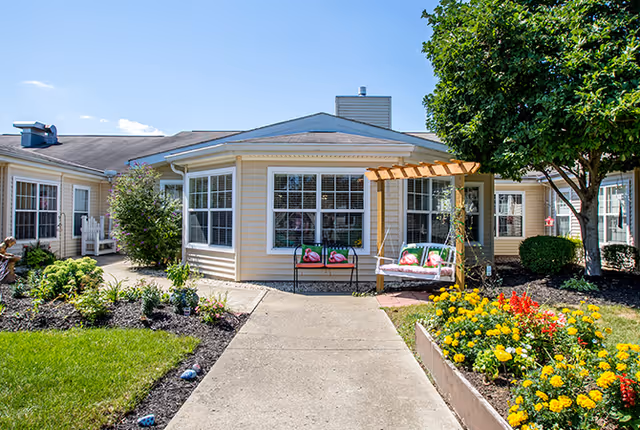 Exterior view of a single-story senior living facility building with beige siding and white trim. A concrete pathway leads to a seating area with a wooden pergola and cushioned bench. The surrounding garden beds are filled with colorful flowers and greenery, and a large tree provides shade on the right side.