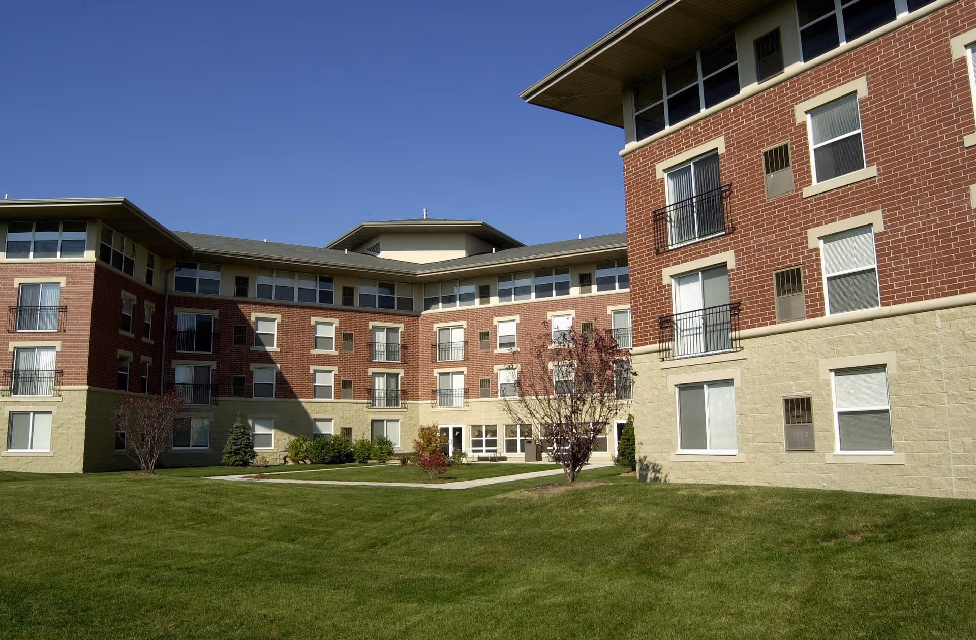 Exterior view of a multi-story senior living facility building with red brick and beige stone facade, surrounded by a well-maintained green lawn and small trees under a clear blue sky.