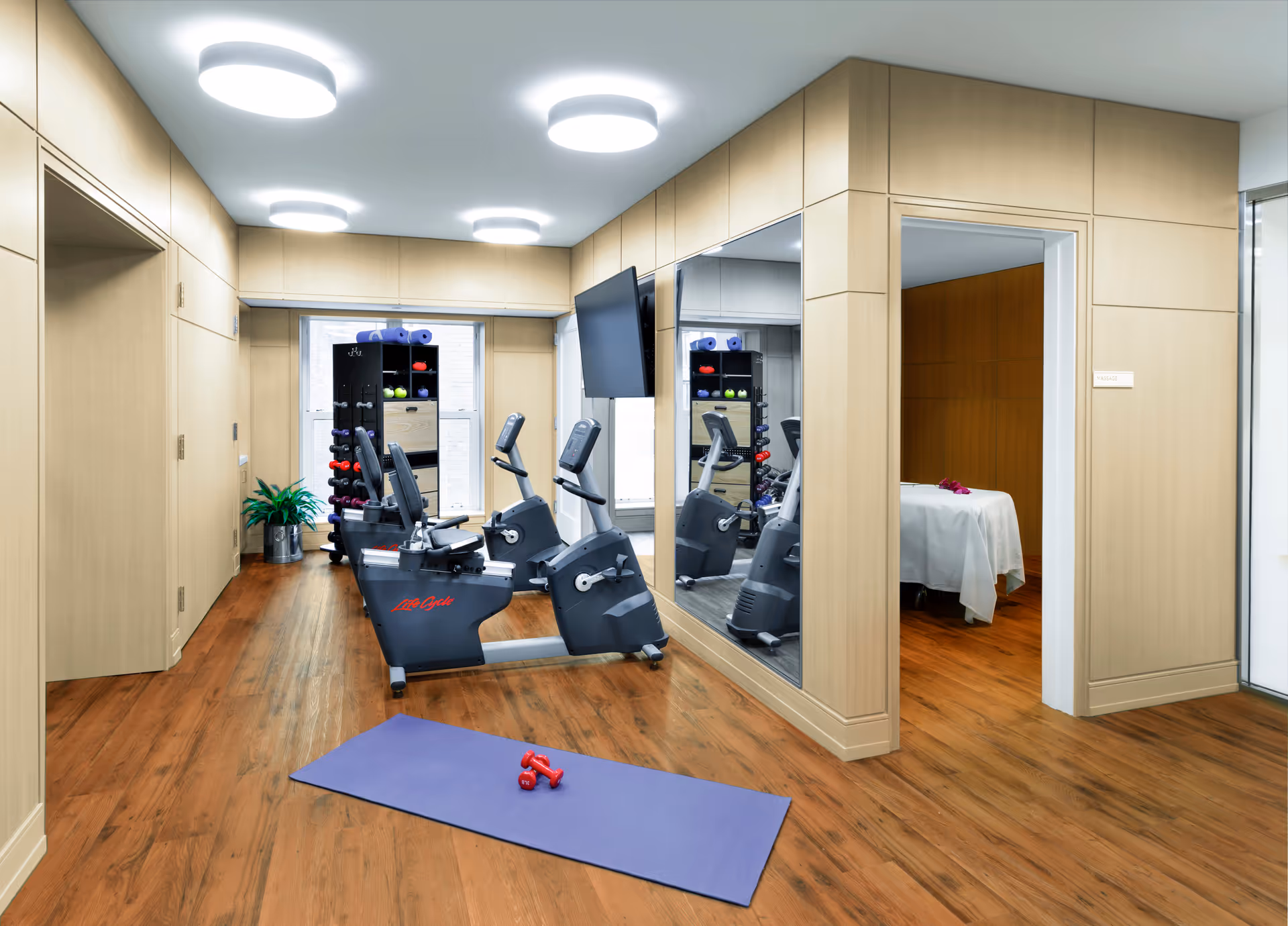 A modern fitness room with two stationary exercise bikes, a rack of colorful dumbbells and yoga mats, a purple exercise mat with two red dumbbells on it, and a large mirror on the wall. Adjacent to the fitness room is a massage room with a massage table covered in a white sheet and a small bouquet of flowers on top. The room has wooden flooring and light wood panel walls with bright ceiling lights.