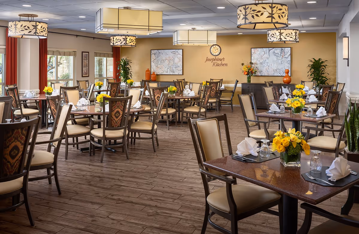A spacious and well-lit dining room with multiple tables and chairs arranged neatly. Each table is set with folded white napkins, glasses, and small vases of yellow flowers. The room features wooden flooring, large windows with red curtains, decorative hanging light fixtures, and wall art. A sign on the back wall reads 'Josephine's Kitchen'.