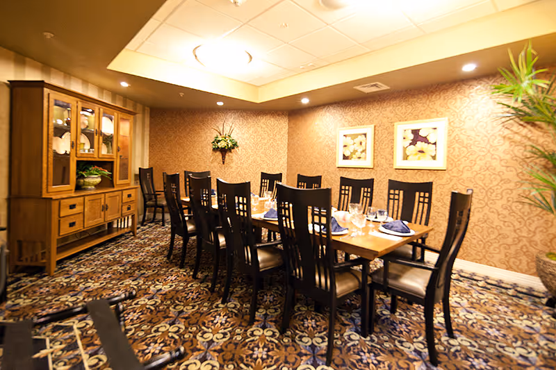 A formal dining room with a long wooden table set for twelve people with black chairs. The table is set with glasses, plates, and blue napkins. The room has patterned carpet, wallpaper with a subtle design, two framed floral pictures on the wall, a wooden cabinet with glass doors displaying dishes, and a potted plant in the corner.