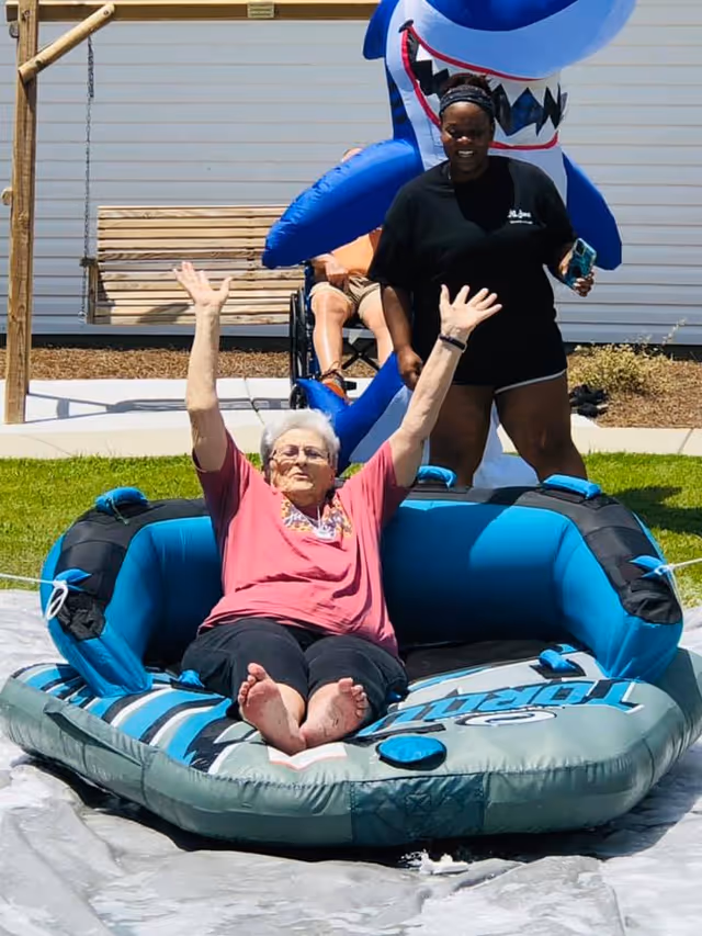 An elderly woman sitting on an inflatable water tube with her arms raised, smiling outdoors on a sunny day. Behind her, a younger woman stands smiling, and a person in a wheelchair is partially visible. There is a wooden swing and a large inflatable shark in the background.