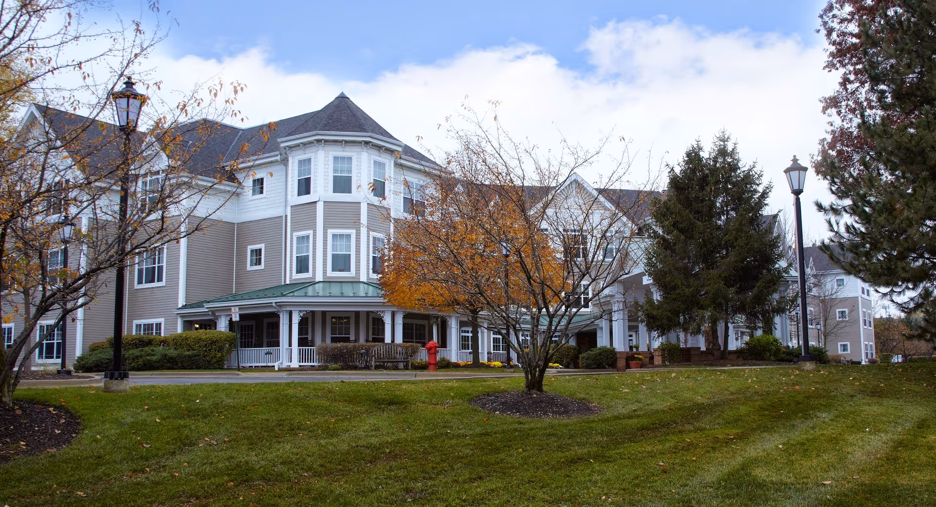 Exterior view of a multi-story senior living facility building with beige and white siding, surrounded by trees with autumn foliage and a well-maintained lawn under a partly cloudy sky.