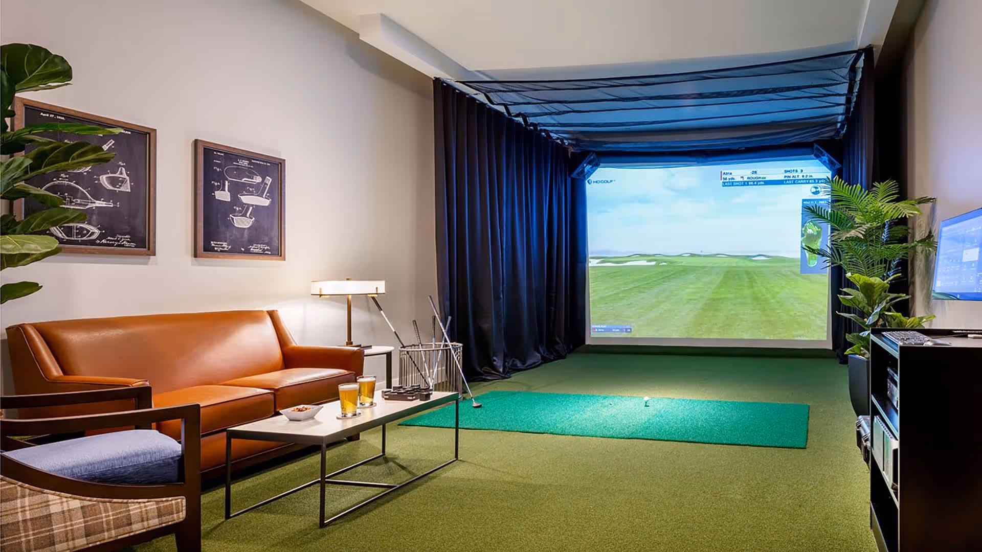 Indoor golf simulator room with a large screen displaying a golf course. The room has green carpet flooring, a brown leather sofa, a wooden armchair with a blue cushion, a small white coffee table with two glasses of beer and a bowl of snacks, golf clubs in a stand, framed golf-themed artwork on the wall, and potted plants.