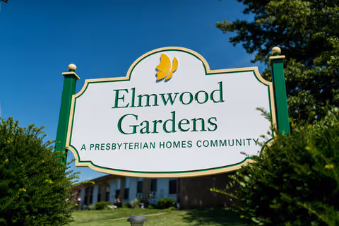 A white and green sign with a yellow butterfly logo that reads 'Elmwood Gardens A Presbyterian Homes Community' set outdoors with bushes and a building in the background under a clear blue sky.