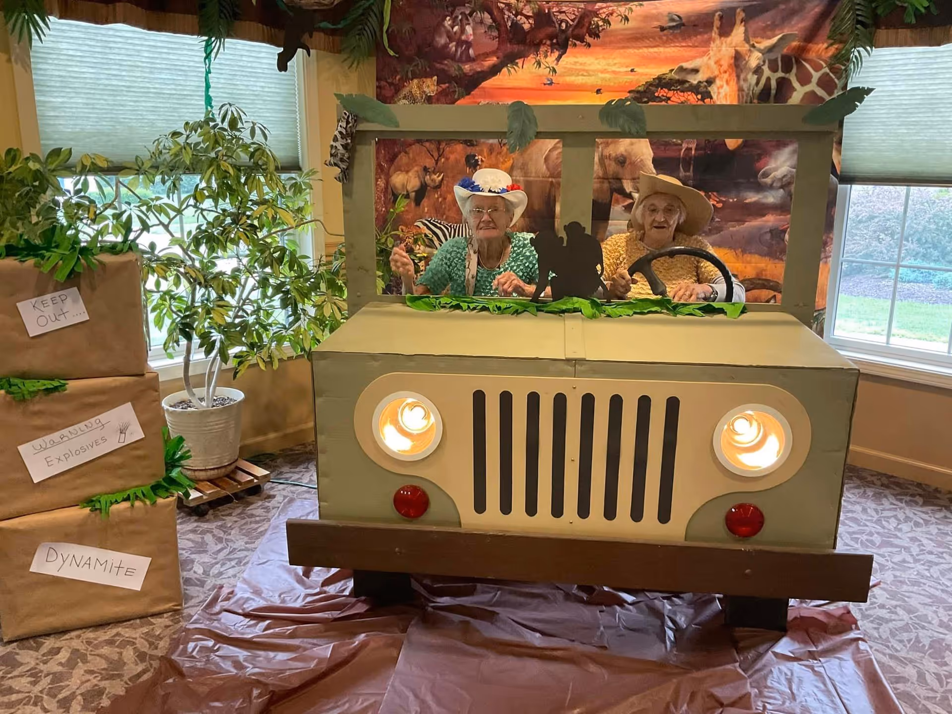 Two elderly women wearing hats sit behind a cardboard cutout designed to look like the front of a jeep. The background features a mural with various wild animals such as giraffes, elephants, and zebras. To the left, there are three large boxes labeled 'KEEP OUT...', 'WARNING Explosives', and 'DYNAMITE'. The setting appears to be indoors with windows showing greenery outside.