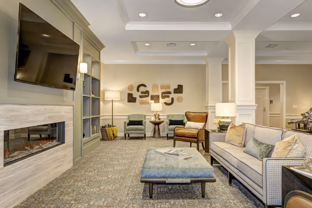 A cozy living room area in a senior living facility featuring a mounted flat-screen TV above a modern electric fireplace, a patterned sofa with multiple cushions, a large upholstered ottoman with books on top, two green armchairs, a brown leather armchair, a floor lamp, table lamps, and abstract wall art. The room has neutral tones with soft lighting and carpeted flooring.