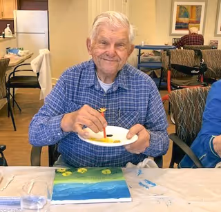 An elderly man wearing a blue checkered shirt is sitting at a table in a communal room, smiling while painting on a canvas. The table is covered with a white cloth, and there are painting supplies and a glass of water in front of him. In the background, another person is seated at a table with a walker nearby.