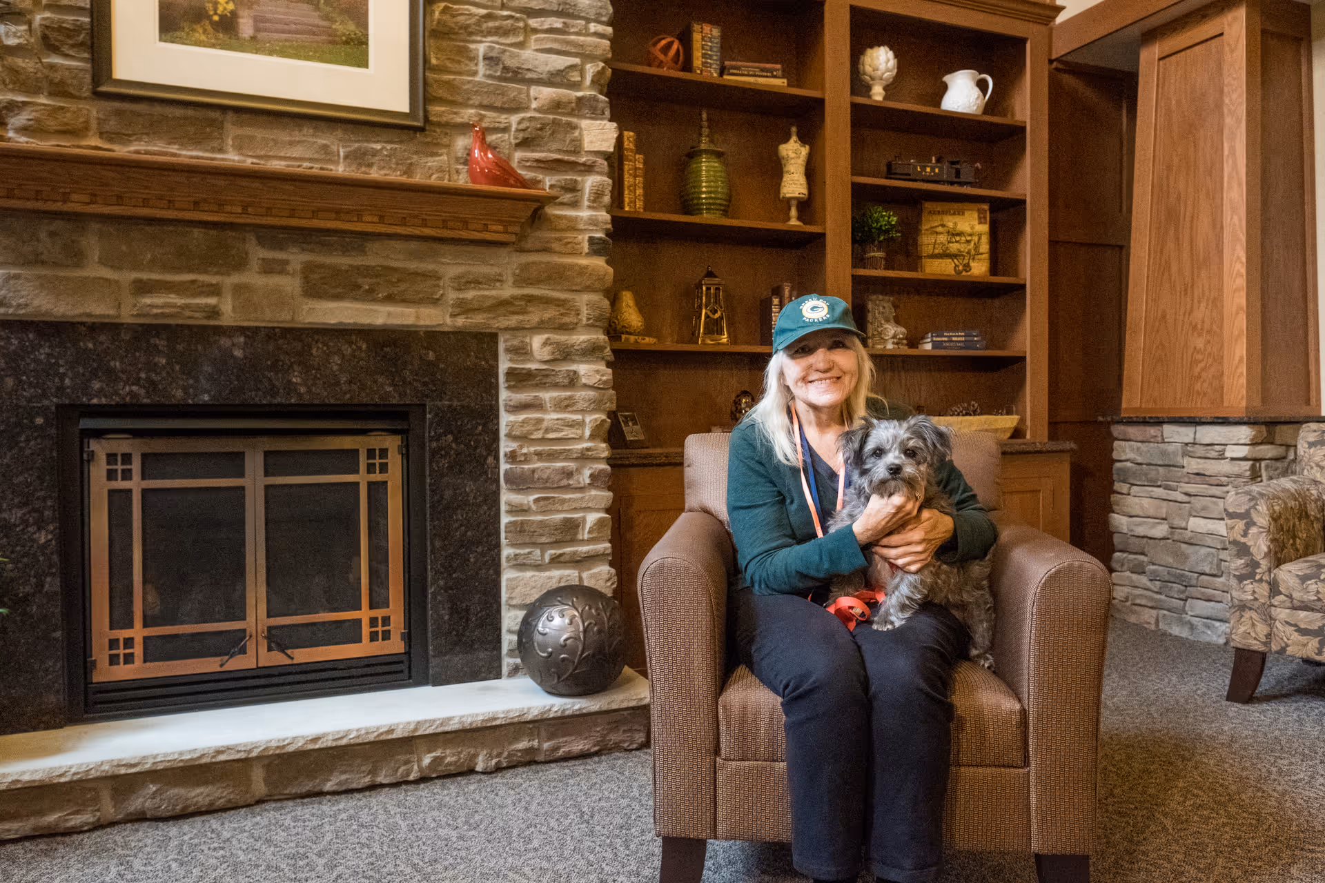 A smiling woman wearing a green cap and jacket sits in a brown armchair in a cozy room with a stone fireplace and wooden shelves. She is holding a small gray dog on her lap. The room has warm lighting and decorative items on the shelves behind her.