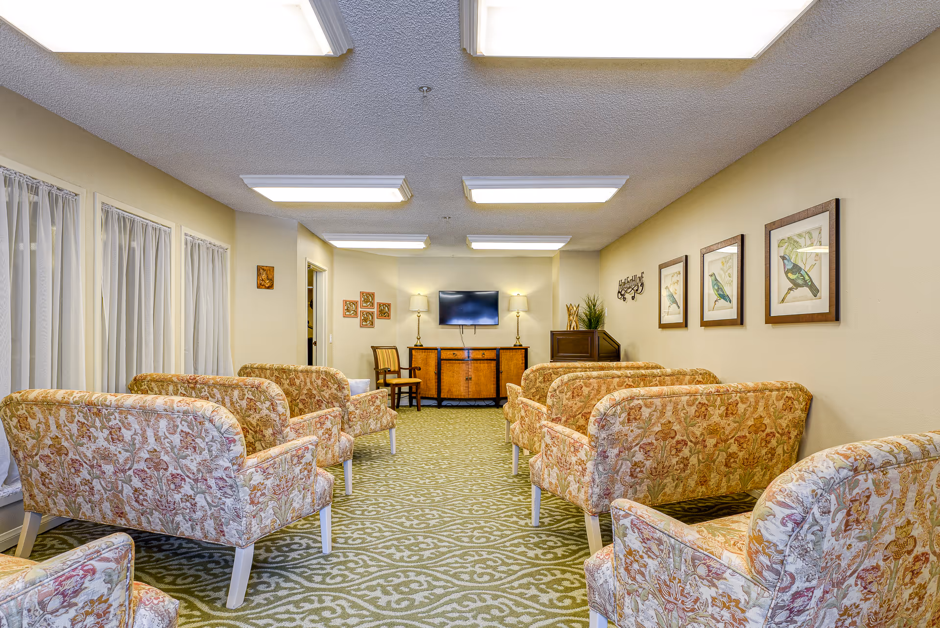 A senior living facility common room with floral patterned armchairs arranged in rows facing a wall-mounted flat screen TV. The room has a green patterned carpet, beige walls decorated with framed bird artwork, and two table lamps on a wooden cabinet beneath the TV. There are windows with sheer white curtains on the left side.
