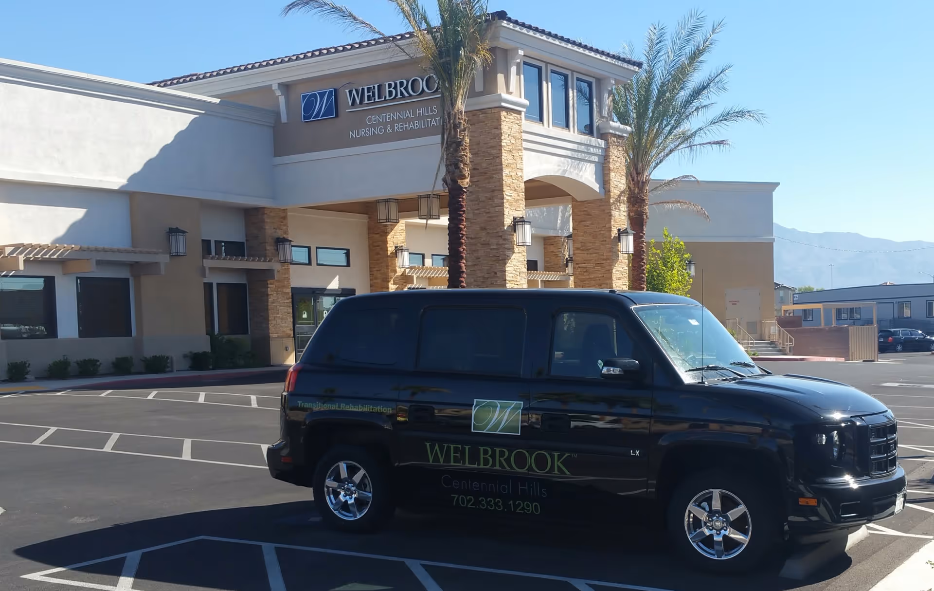 Exterior view of a nursing and rehabilitation facility named Welbrook Centennial Hills with a black Welbrook branded vehicle parked in front. The building has stone pillars, palm trees, and a clear sky in the background.