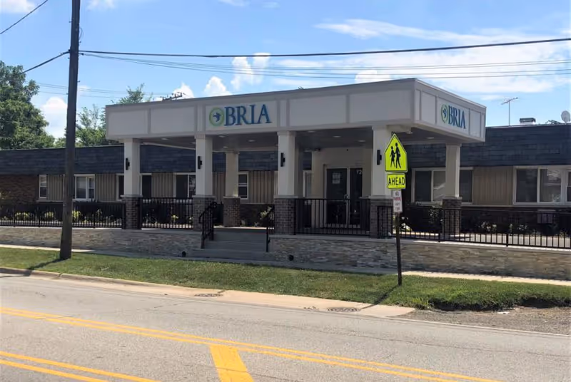 Exterior front view of the BRIA of Chicago Heights facility showing a single-story building with a covered entrance supported by columns. The building has a sign with the BRIA logo and name. There is a pedestrian crossing sign in front of the entrance and a road with yellow lines in the foreground.