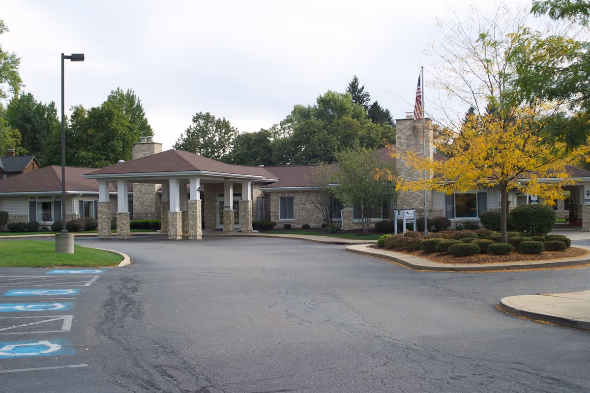 Exterior view of a single-story senior living facility building with a covered entrance supported by brick columns. There is a driveway leading up to the entrance, surrounded by landscaped bushes and trees, one of which has yellow autumn leaves. An American flag is visible near the entrance, and there are several parking spaces, including handicapped spots, in the foreground.