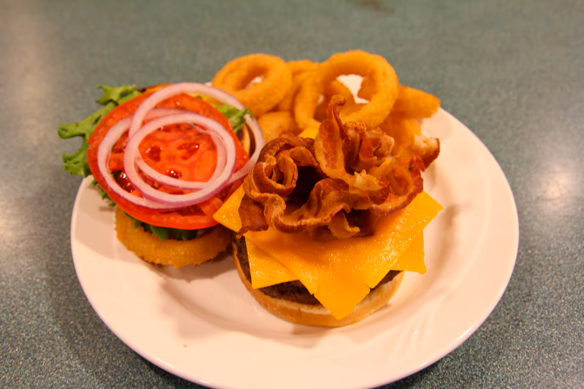 A plate with a bacon cheeseburger topped with cheddar cheese and crispy bacon, served with onion rings and garnished with lettuce, tomato slices, and red onion rings on the side.