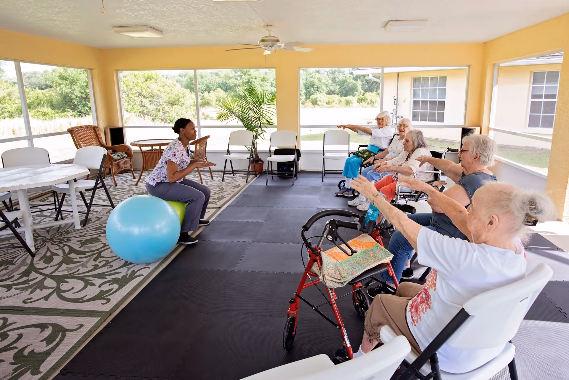 A group of elderly women seated in chairs in a bright, enclosed patio area participating in a seated exercise session led by a caregiver sitting on an exercise ball. The room has large windows showing greenery outside, a ceiling fan, and a patterned rug on one side.