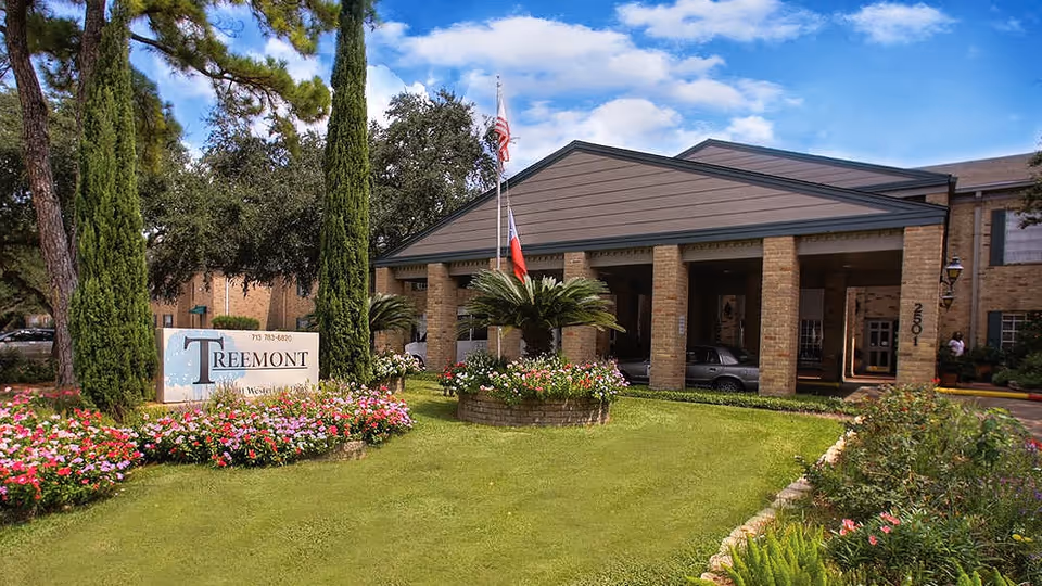 Exterior view of Treemont Senior Living facility showing a brick building with a covered entrance supported by columns. The foreground features a well-maintained lawn with colorful flower beds and tall trees. Two flagpoles with American and Texas flags are visible near the entrance.