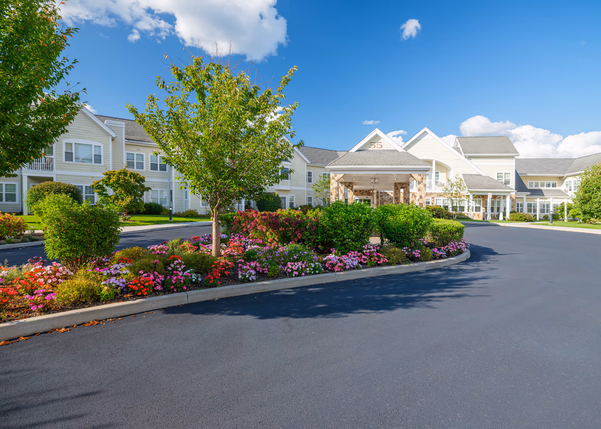 Exterior view of The Knolls, A Bethel Community senior living facility on a sunny day with a clear blue sky. The building is light-colored with multiple windows and a covered entrance marked with the number 55. In front of the building, there is a curved driveway bordered by well-maintained flower beds and green shrubs and trees.