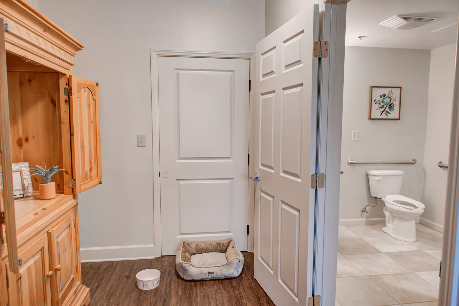 Interior view of a room with a wooden cabinet on the left, a pet bed and a pet food bowl on the floor in front of a closed white door. To the right, an open door reveals a bathroom with a toilet, grab bars, tiled floor, and a framed floral picture on the wall.