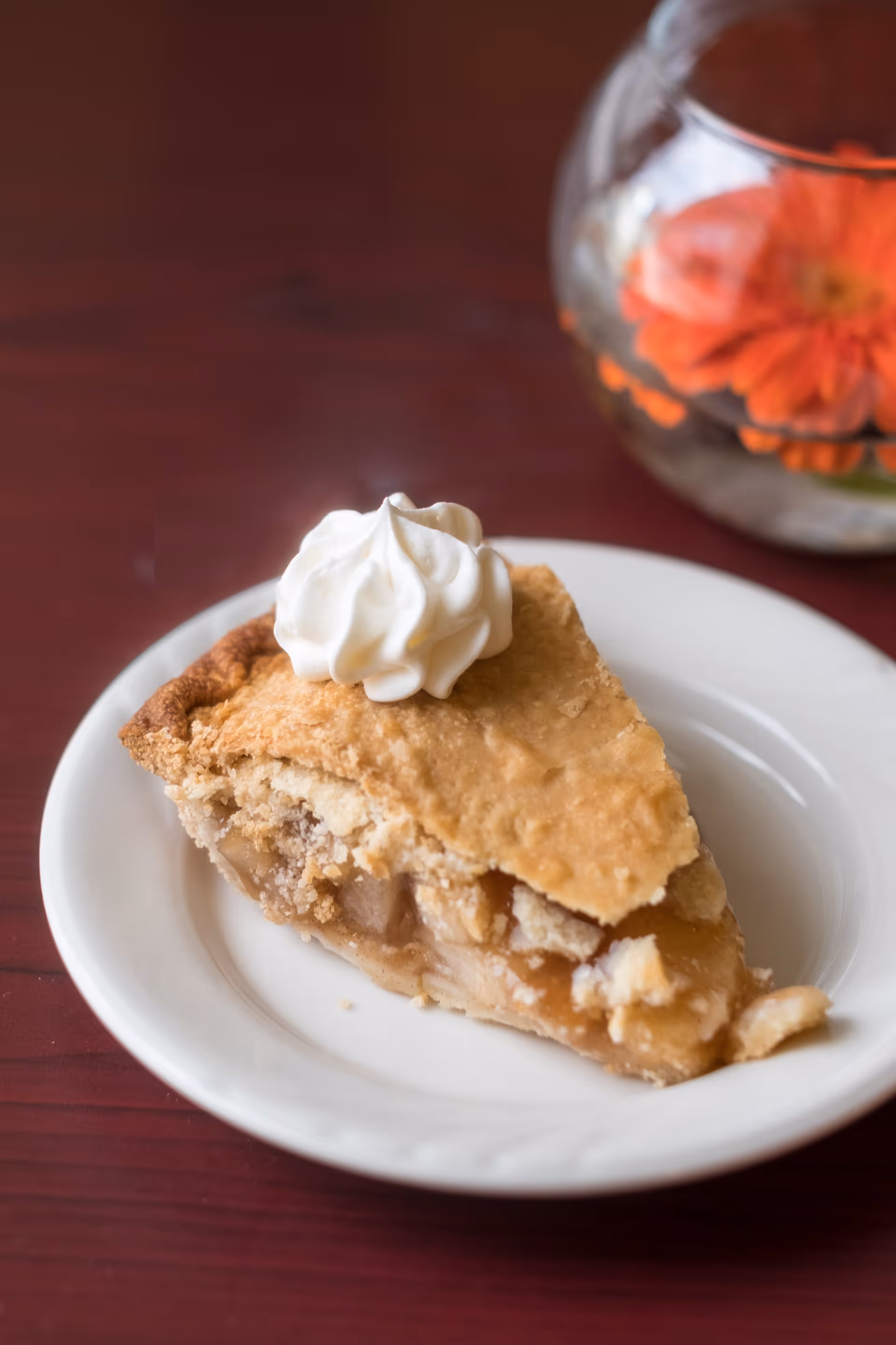A slice of apple pie topped with a dollop of whipped cream on a white plate, placed on a dark wooden table with a glass bowl containing an orange flower in the background.