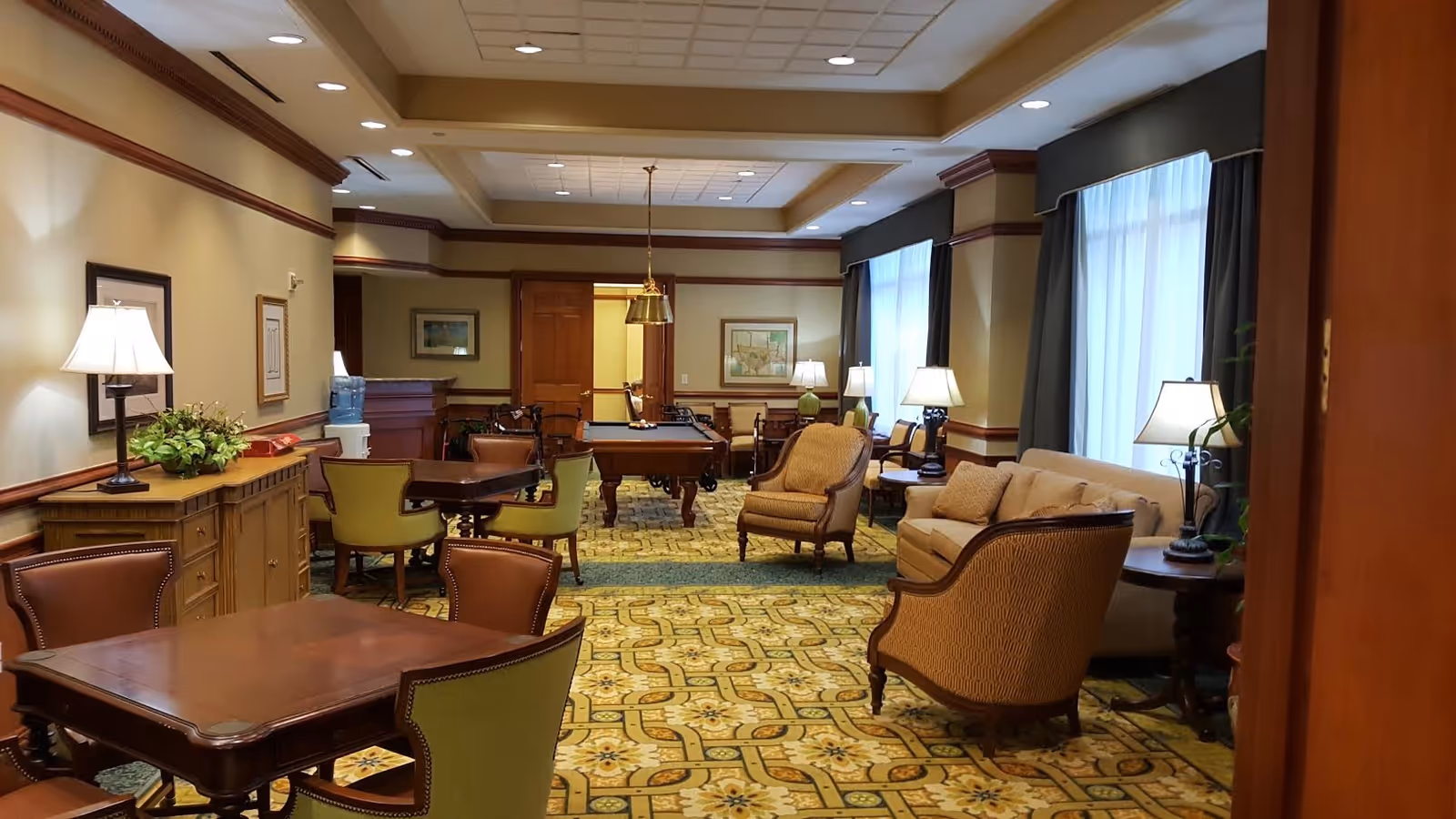 A spacious, well-lit common area with multiple seating options including armchairs, sofas, and tables with chairs. The room features large windows with curtains, several table lamps, framed artwork on the walls, and a pool table in the background. The carpet has a patterned design and the walls are painted in warm tones with wooden trim.