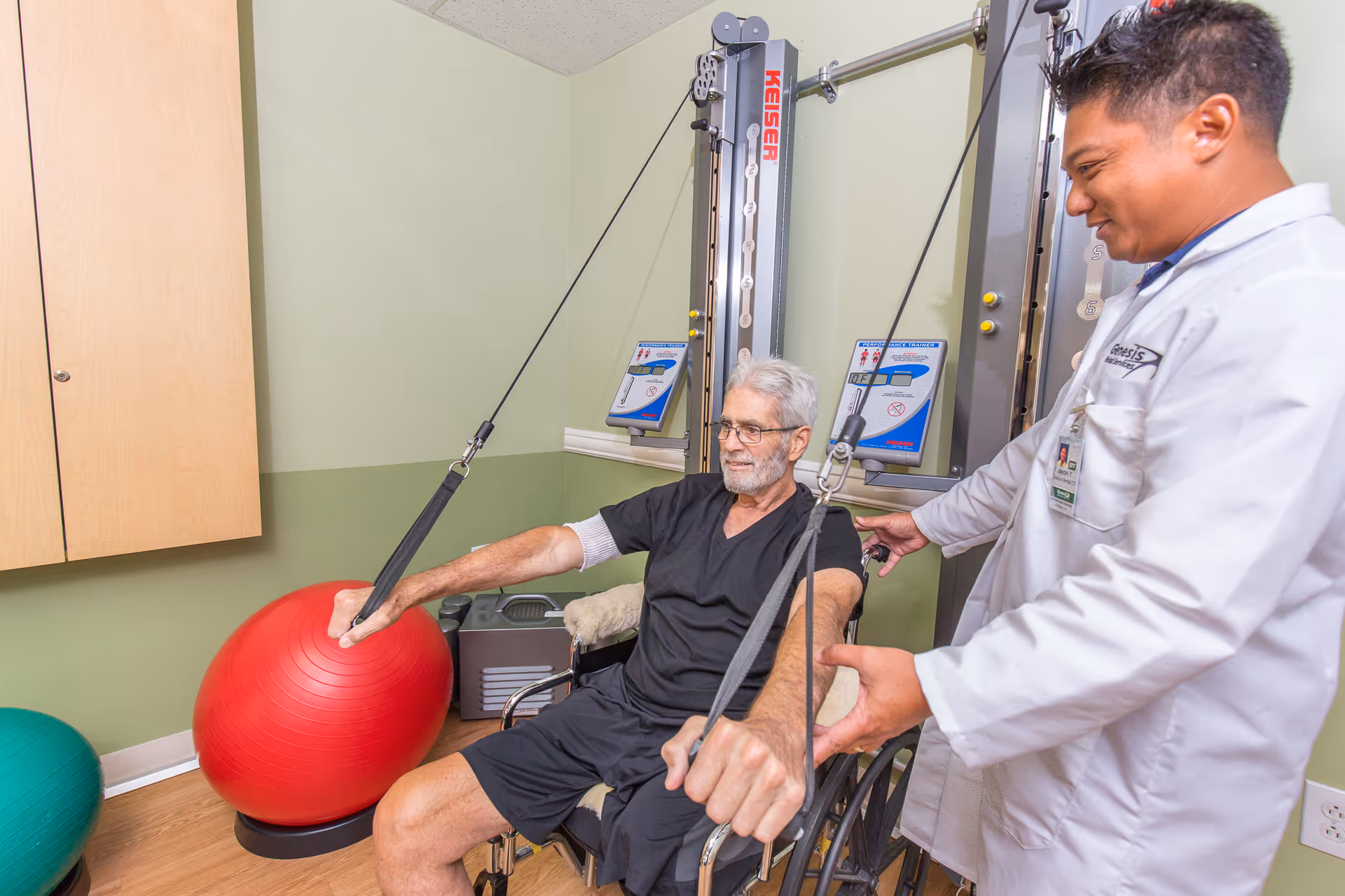 An elderly man in a wheelchair is exercising his arms using resistance cables attached to a Keiser exercise machine, assisted by a healthcare professional in a white coat in a therapy room with light green walls and wooden flooring.
