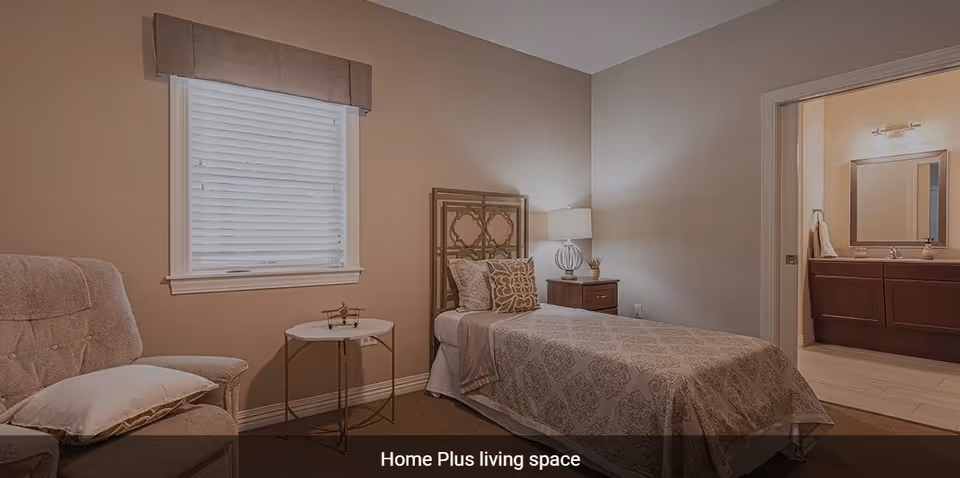 A cozy bedroom with a single bed covered in patterned bedding, a cushioned armchair with a pillow, a small round side table, and a window with blinds and a valance. There is a nightstand with a lamp next to the bed, and an open doorway leading to a bathroom with a vanity and mirror.
