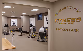 Interior view of the Belmont Village Senior Living Lincoln Park fitness center showing exercise equipment including stationary bikes and elliptical machines, with framed artwork on the walls and a sign reading 'Belmont Village Fitness Center Lincoln Park'.