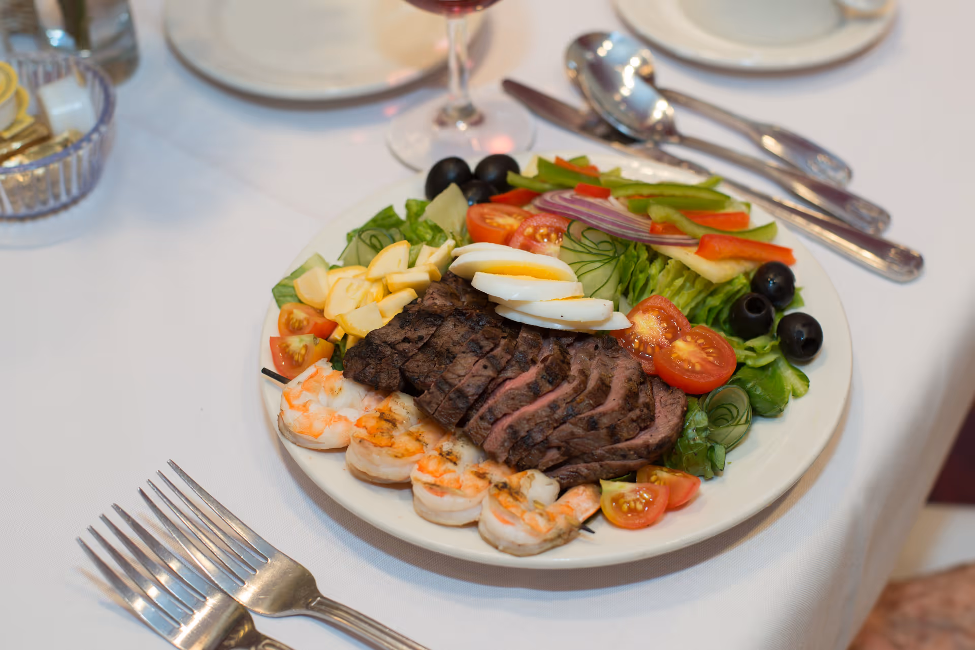 A plate of food on a white tablecloth featuring grilled shrimp skewers, sliced grilled steak, hard-boiled egg slices, cherry tomatoes, black olives, cucumber slices, red and green bell pepper strips, and lettuce. There are forks, spoons, and a glass of water visible on the table.