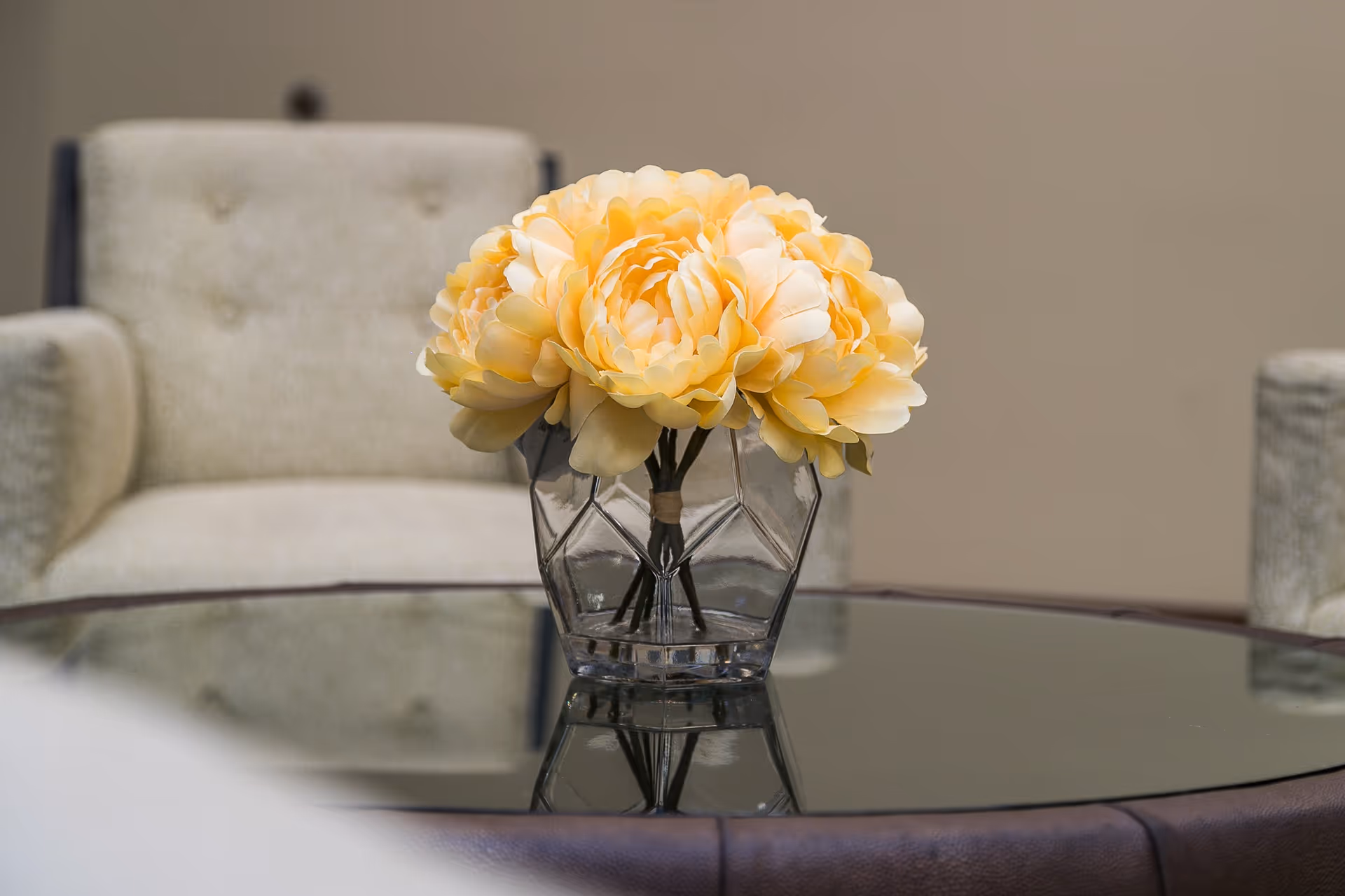 A clear glass vase with a geometric design holding a bouquet of light yellow flowers sits on a reflective glass coffee table. In the background, there are two beige upholstered armchairs against a plain beige wall.