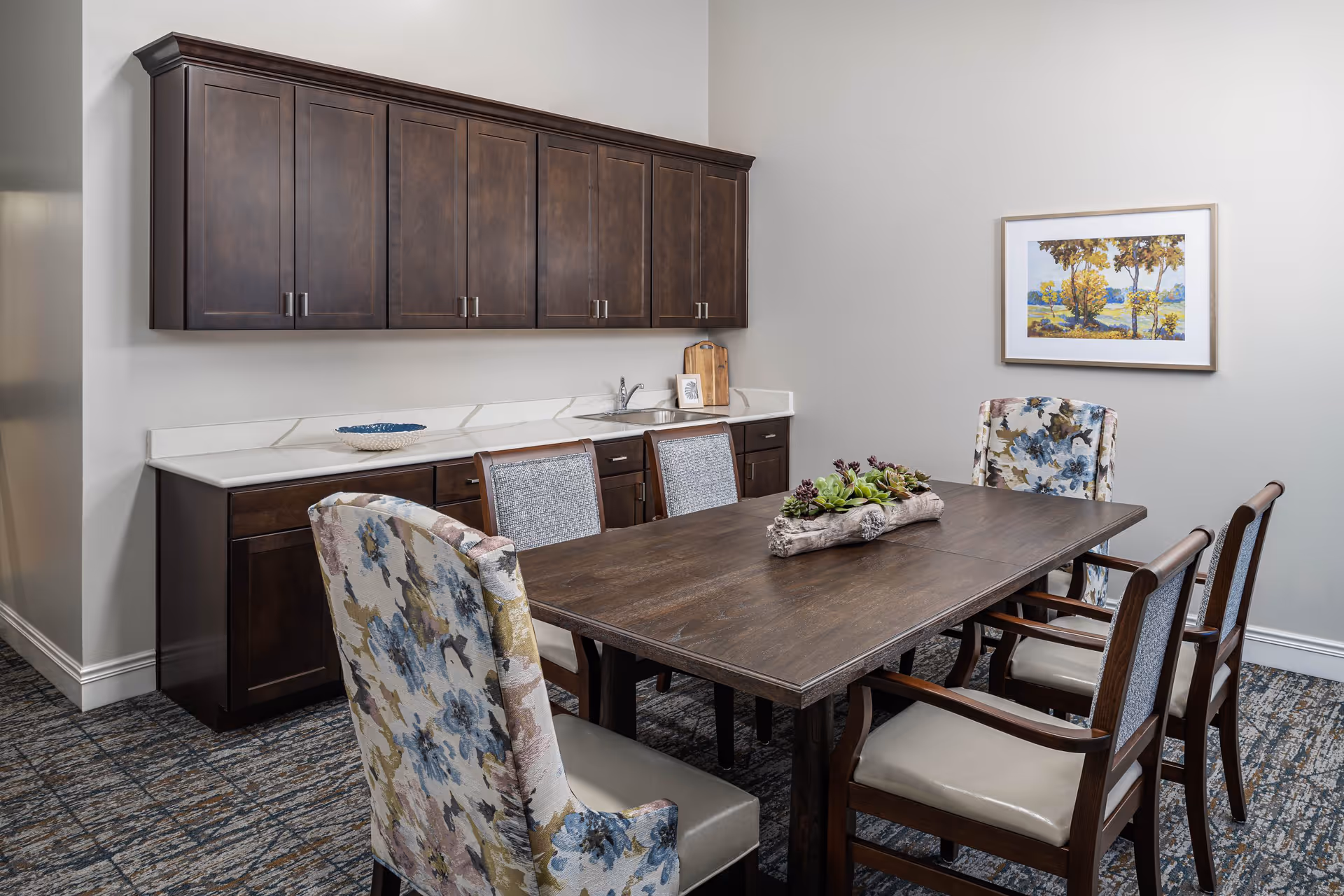A dining area with a dark wooden table surrounded by six chairs, two of which have floral upholstery and the others have gray fabric backs and cream-colored seats. Behind the table is a countertop with a small sink and dark wooden cabinets above and below. A framed landscape painting hangs on the light gray wall.