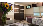 A well-lit bedroom in a healthcare facility featuring two beds with floral bedspreads, a dark wood wardrobe, a small wall-mounted television, and a window with blinds partially drawn. A vase with sunflowers and other flowers is placed on a table in the foreground.