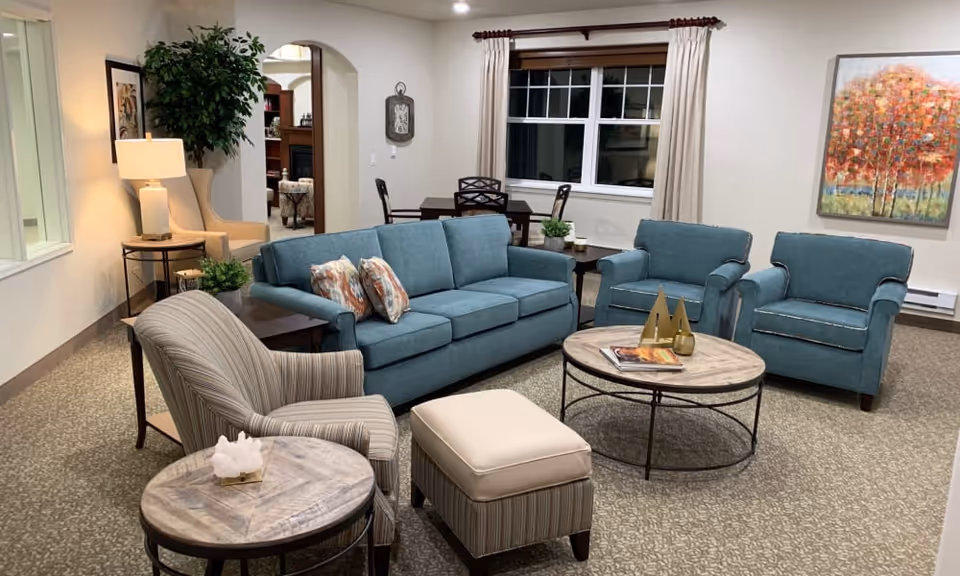 A cozy living room with a blue sofa, two matching blue armchairs, a striped armchair, and a striped ottoman. There are two round wooden coffee tables, one with decorative items and magazines. The room has a large window with beige curtains, a colorful painting on the wall, a floor lamp, and a potted plant near an archway leading to another room.