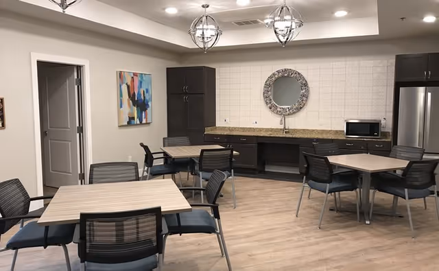 A communal dining area with four square tables, each surrounded by four chairs with black mesh backs and blue cushions. The room features light wood flooring, beige walls, and a kitchenette along the back wall with dark cabinets, a granite countertop, a microwave, and a stainless steel refrigerator. Above the countertop is a round decorative mirror. Two modern pendant lights hang from the ceiling.