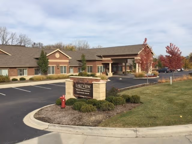 Exterior view of Lakeview Memory Care facility showing a single-story brick building with a covered entrance, a parking lot, landscaped bushes, and trees with autumn foliage under a partly cloudy sky.