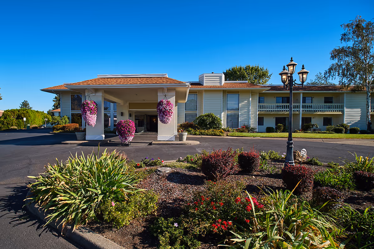 Exterior view of Northridge Senior Living building with a covered entrance, hanging flower baskets, landscaped garden with various plants and flowers, and a vintage-style street lamp under a clear blue sky.
