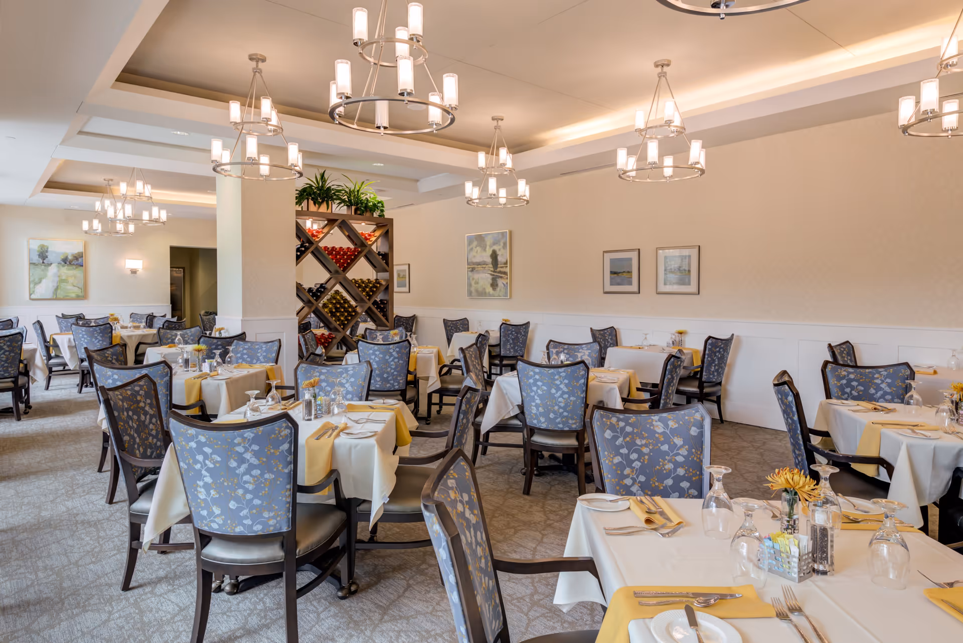 A spacious dining room with multiple tables covered in white tablecloths and yellow napkins. Each table is set with plates, silverware, glasses, and small floral centerpieces. The chairs have blue floral upholstery. The room features soft lighting from modern chandeliers and framed artwork on the walls. A wine rack with bottles and plants is visible in the background.