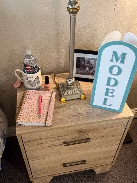 A wooden nightstand with two drawers holding a table lamp, a framed black and white photo, a white mug with a water bottle inside labeled 'Blessed Mom', a bottle of pink nail polish, a pink pen on a pink and gold polka dot notebook, and a blue and white sign that reads 'MODEL'.