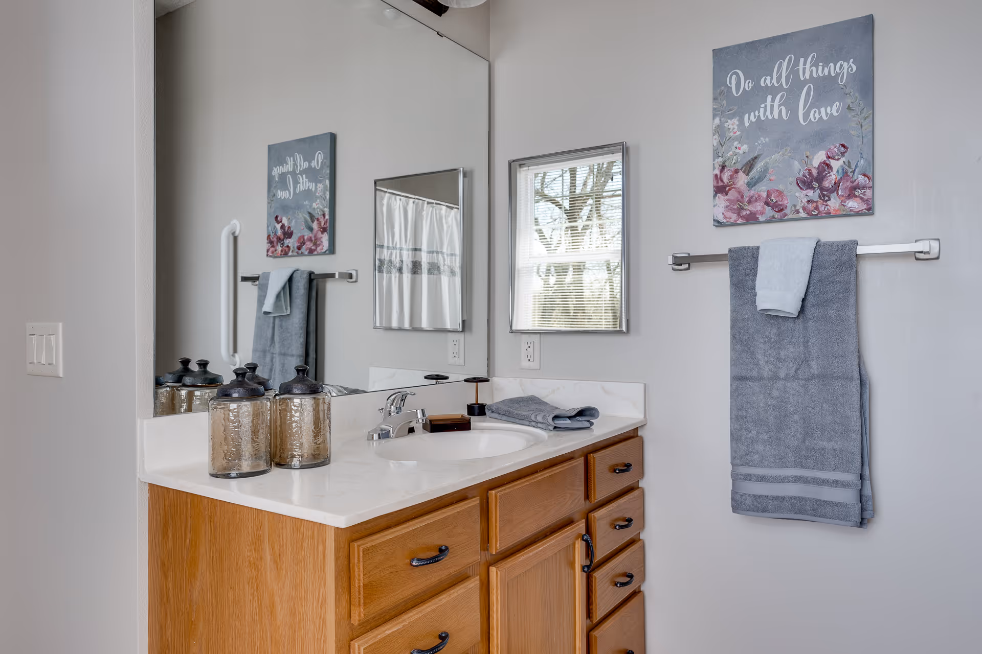 A bathroom vanity with a wooden cabinet and a white countertop featuring a sink and faucet. Two decorative glass jars are placed on the countertop. Above the vanity is a large mirror reflecting a small window with blinds and a shower curtain. On the wall next to the mirror, there is a towel rack holding two gray towels and a decorative sign that reads 'Do all things with love' with floral designs.