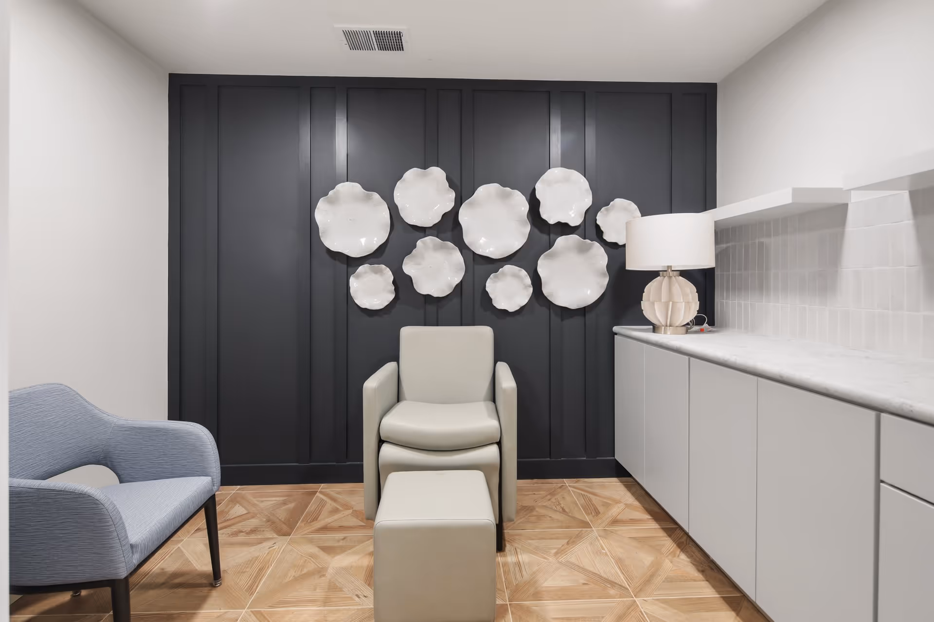 A modern interior room with a light gray armchair and matching ottoman centered against a dark paneled wall decorated with white abstract ceramic wall art. To the left is a light blue upholstered chair, and to the right is a white countertop with cabinets below and a decorative table lamp on top. The floor has a wood-patterned tile design.